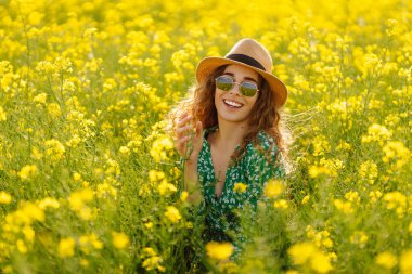 Mutlu kadın eğleniyor ve kolza tohumu tarlasında yürüyor. Parlak elbiseli ve şapkalı güzel bir kadın güneşli havanın tadını çıkarıyor. Doğa kavramı, rahatlama..