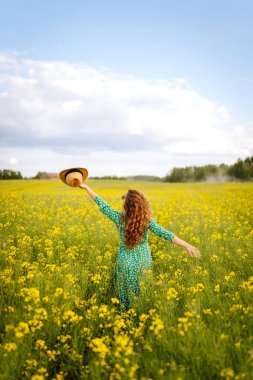 Mutlu kadın eğleniyor ve kolza tohumu tarlasında yürüyor. Parlak elbiseli ve şapkalı güzel bir kadın güneşli havanın tadını çıkarıyor. Doğa kavramı, rahatlama..