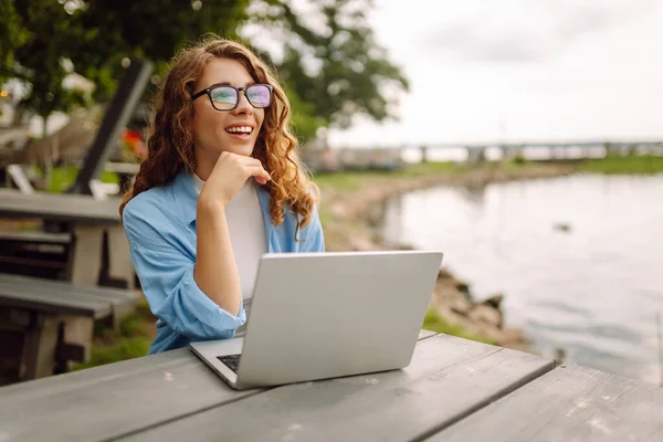 Cheerful woman in casual clothes sitting at a table by the lake with a laptop. Cute woman working with laptop outdoors. Freelancing, technology concept.