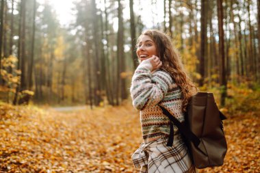 Sırt çantalı genç ve güzel bir turist sonbahar ormanında eğleniyor, yapraklarını döküyor. Kadın ormanda renkli bir patika boyunca yürüyor. Özgürlük, seyahat.