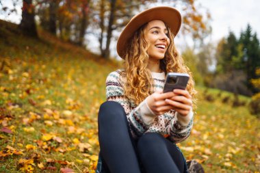 Sonbahar parkında, sarı yaprakların arasında telefonu olan, şapkalı, kıvırcık bir kadın. Açık havada akıllı telefon kullanan gülümseyen bir kadın. Tatil, hafta sonu, teknoloji kavramı.