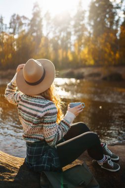 Şapkalı genç bir kadın gezgin nehir kıyısında sonbahar doğasının tadını çıkarıyor, termostan sıcak bir içecek içiyor. Güzel bir kadın çay içiyor, gölün kenarında oturuyor..