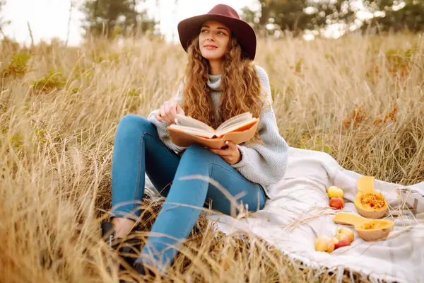 穿着舒适的毛衣和帽子的年轻女人喜欢大自然 一边看书 一边坐在秋天公园的毛毯上 一个漂亮的女人正在户外野餐 — 图库照片