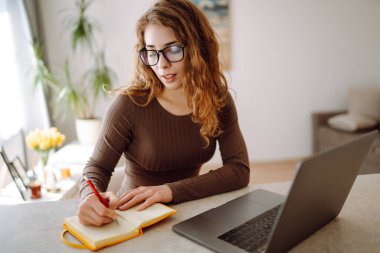 Odaklanmış genç bir kadın elinde dizüstü bilgisayar ve parlak bir not defteriyle masada oturur, çalışır, ekrana bakar, masaya yazar. Serbest çalışma, tatil, eğitim kavramı.