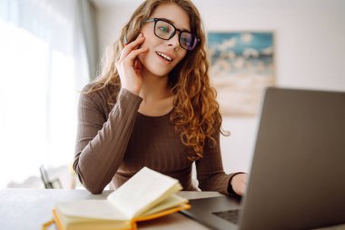 Odaklanmış genç bir kadın elinde dizüstü bilgisayar ve parlak bir not defteriyle masada oturur, çalışır, ekrana bakar, masaya yazar. Serbest çalışma, tatil, eğitim kavramı.