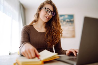 Odaklanmış genç bir kadın elinde dizüstü bilgisayar ve parlak bir not defteriyle masada oturur, çalışır, ekrana bakar, masaya yazar. Serbest çalışma, tatil, eğitim kavramı.