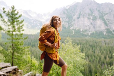 Yürüyüş yolunda parlak bir sırt çantasıyla gülümseyen bir gezgin. Uçurumun tepesinde güzel bir kadın dağ manzarasının tadını çıkarıyor. Etkin yaşam biçimi.