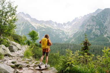 Yürüyüş yolunda parlak bir sırt çantasıyla gülümseyen bir gezgin. Uçurumun tepesinde güzel bir kadın dağ manzarasının tadını çıkarıyor. Etkin yaşam biçimi.