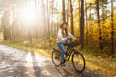 Şık bir süveter ve şapka giyen genç bir kadın bisiklet sürüyor, özgürlüğü hissediyor, ormanda sonbahar havasının tadını çıkarıyor. Açık hava portresi. Etkin yaşam biçimi.