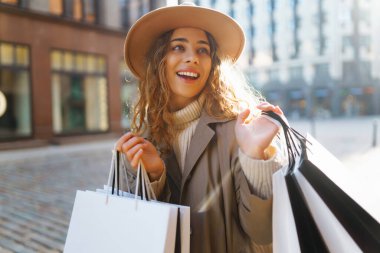 Paltolu ve şapkalı şık bir kadın güneşli bir havada alışveriş merkezinin yakınında yürüyor. Güzel bir kadın Avrupa sokaklarından zevk alır. Tüketim, alışveriş, alışveriş kavramı.