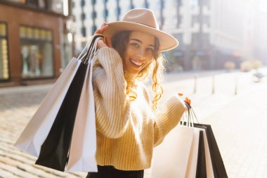 Paltolu ve şapkalı şık bir kadın güneşli bir havada alışveriş merkezinin yakınında yürüyor. Güzel bir kadın Avrupa sokaklarından zevk alır. Tüketim, alışveriş, alışveriş kavramı.