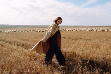 Bir tarlada duran ceketli genç bir kadın kuru otların arasında, doğanın tadını çıkarıyor. Şehir tarzı ve sokak modası. Doğa, moda, özgürlük kavramı. Yaşam biçimi.