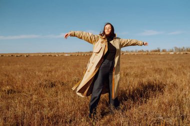 Bir tarlada duran ceketli genç bir kadın kuru otların arasında, doğanın tadını çıkarıyor. Şehir tarzı ve sokak modası. Doğa, moda, özgürlük kavramı. Yaşam biçimi.