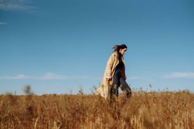 Bir tarlada duran ceketli genç bir kadın kuru otların arasında, doğanın tadını çıkarıyor. Şehir tarzı ve sokak modası. Doğa, moda, özgürlük kavramı. Yaşam biçimi.