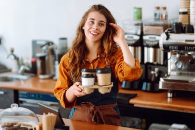 Güzel bir kahve dükkanı sahibi elinde sıcak içecek dolu kağıt bardaklar tutuyor. Genç bayan barista tezgahın arkasında çalışıyor. Paket yemek..