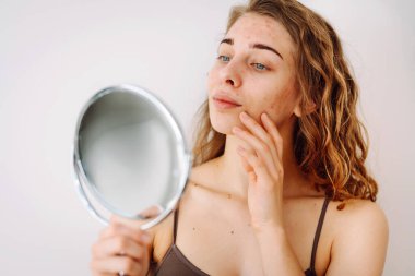 Yüzünde isilik olan tatlı bir kadın küçük bir aynaya bakıyor. Güzel bir kadın sorunlu cildini inceliyor. Doğal güzellik. Kozmetoloji, dikkat.