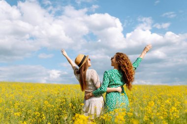 İki neşeli kız arkadaş, kolza tohumu tarlasında yürürken harika vakit geçiriyorlar. Yürüme, aktif yaşam tarzı, tatiller.