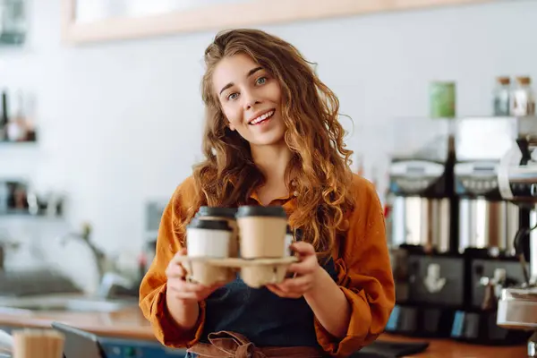 A beautiful coffee shop owner holds paper cups with hot takeaway drinks in her hand. Young female barista works behind the counter. Takeaway food.