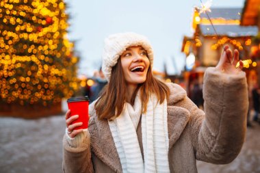 Kış tarzı kıyafetler giymiş genç bir kadın, festival caddesi pazarında poz veriyor. Kış anlarından hoşlanan kadın. Güzel ışık süslemeleri. Noel festivali, kış tatili konsepti.. 