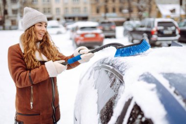 Karı temizleme zamanı! Genç bir kadın bir fırçayla arabadan karı temizliyor. Kış arabası konsepti.