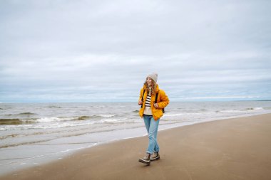 Sırt çantalı, sarı ceketli genç ve güzel bir turist kadın deniz kenarında hayatın tadını çıkarıyor. Seyahat, yaşam tarzı, macera.