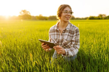 Genç kadın tarım uzmanı yeşil buğday tarlasında modern dijital tablet üzerinde çalışıyor. Yaşlı bir çiftçi, tablet kullanan genç buğday filizlerinin kalitesini kontrol ediyor..