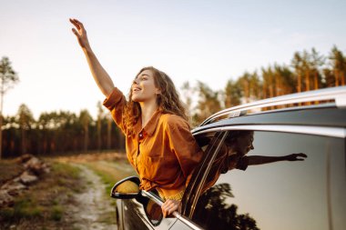 Genç bir kadın, güneşli bir ormanda arabanın camından sarkan özgürlüğü hissediyor. Bir gezgin araba penceresinden güneşli bir günün tadını çıkarır. Seyahat konsepti. Etkin yaşam biçimi.