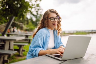 Göl kenarındaki bir kafede dizüstü bilgisayarla çalışan günlük kıyafetli mutlu bir kadın. Doğanın tadını çıkaran serbest çalışan genç bir kadın. Tenoloji konsepti, çalışma günü.