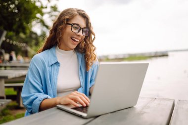 Göl kenarındaki bir kafede dizüstü bilgisayarla çalışan günlük kıyafetli mutlu bir kadın. Doğanın tadını çıkaran serbest çalışan genç bir kadın. Tenoloji konsepti, çalışma günü.