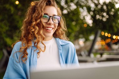 Göl kenarındaki bir kafede dizüstü bilgisayarla çalışan günlük kıyafetli mutlu bir kadın. Doğanın tadını çıkaran serbest çalışan genç bir kadın. Tenoloji konsepti, çalışma günü.