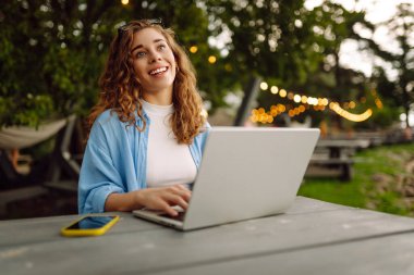 Göl kenarındaki bir kafede dizüstü bilgisayarla çalışan günlük kıyafetli mutlu bir kadın. Doğanın tadını çıkaran serbest çalışan genç bir kadın. Tenoloji konsepti, çalışma günü.