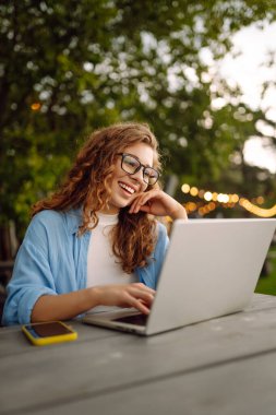 Göl kenarındaki bir kafede dizüstü bilgisayarla çalışan günlük kıyafetli mutlu bir kadın. Doğanın tadını çıkaran serbest çalışan genç bir kadın. Tenoloji konsepti, çalışma günü.