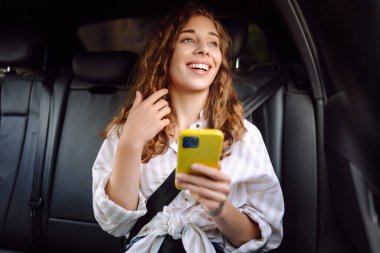 Bir arabanın arka koltuğunda elinde akıllı bir telefonla genç kıvırcık bir kadının portresi. Arabadaki gülümseyen bir kadın telefonu kullanır. Tenoloji kavramı, arabayla seyahat etmek..