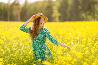 Şapkalı mutlu bir kadın çiçek açan bir kolza tarlasında yürüyor. Güneşli bir günde kolza tohumu tarlasında poz veren güzel bir kadın. Doğa kavramı, rahatlama..