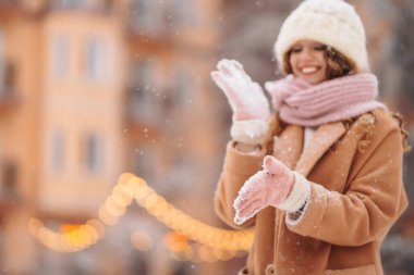 Kış kıyafetleri içinde mutlu bir kadın Noel pazarında yürüyor. Kıvırcık saçlı kadın karlı bir caddede kış tatilinin tadını çıkarıyor. Her yer aydınlık..