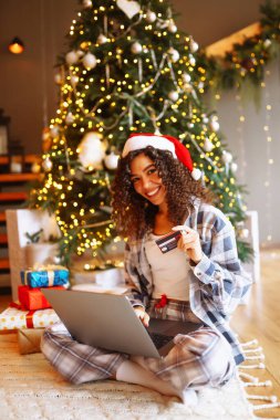 Kıvırcık saçlı, kredi kartı, hediyeleri ve dizüstü bilgisayarı olan gülümseyen bir kadın. Noel ağacının yanındaki evde oturan genç bir kadın internetten alışveriş yapıyor. Alışveriş konsepti, tatil.