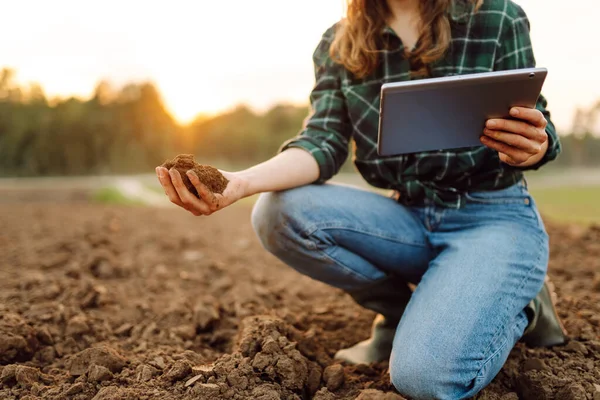 Young woman agronomist checks the quality of the soil before sowing. Farmer woman with a digital tablet holds black soil in her hands. Concept of technology, ecology.