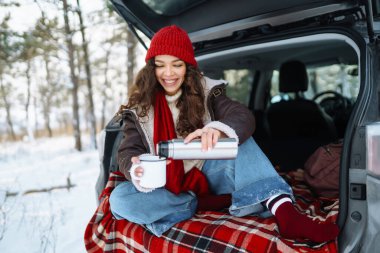 Kırmızı şapkalı genç bir gezgin arabanın bagajında oturur ve termostan sıcak bir içecek içer. Mutlu kadın ormanda karlı manzaranın tadını çıkarıyor. Konfor konsepti, seyahat.