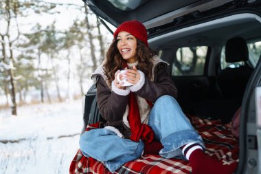 Kırmızı şapkalı genç bir gezgin arabanın bagajında oturur ve termostan sıcak bir içecek içer. Mutlu kadın ormanda karlı manzaranın tadını çıkarıyor. Konfor konsepti, seyahat.
