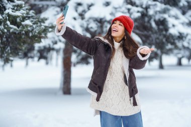 Kırmızı şapkalı, elinde telefon olan mutlu bir kadın kış ormanında selfie çekiyor. Parktaki karlı manzaranın tadını çıkaran tatlı bir kadın. Doğa kavramı, teknoloji.