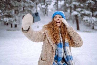Mutlu kadın, karlı bir kış ormanında akıllı telefon kullanarak selfie çekiyor. Blog yazma, seyahat, hafta sonu kavramı. Etkin yaşam biçimi.