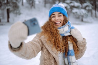 Mutlu kadın, karlı bir kış ormanında akıllı telefon kullanarak selfie çekiyor. Blog yazma, seyahat, hafta sonu kavramı. Etkin yaşam biçimi.