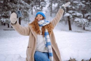 Mutlu kadın, karlı bir kış ormanında akıllı telefon kullanarak selfie çekiyor. Blog yazma, seyahat, hafta sonu kavramı. Etkin yaşam biçimi.