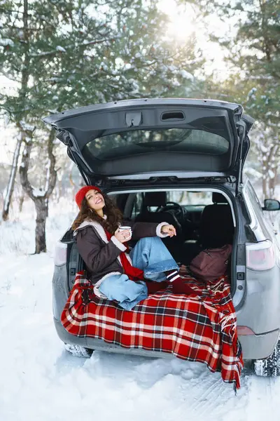 A young traveler in a red hat sits in the trunk of a car and drinks a hot drink from a thermos. Happy woman enjoying snowy landscape in the forest. The concept of comfort, travel.