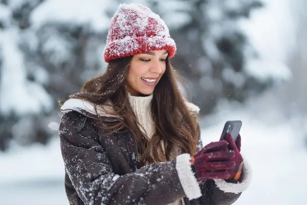 Happy woman in a red hat with a phone in her hands takes a selfie, blogs in the winter forest. Cute woman enjoying the snowy landscape in the park. Concept of nature, technology.
