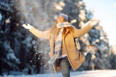 Karla oynamak. Şapkalı ve atkılı güzel bir kadın karla oynuyor, karlı bir kış ormanında eğleniyor. Neşeli kadın güneşli ve soğuk havanın tadını çıkarıyor..