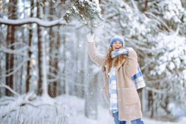 Karla oynamak. Şapkalı ve atkılı güzel bir kadın karla oynuyor, karlı bir kış ormanında eğleniyor. Neşeli kadın güneşli ve soğuk havanın tadını çıkarıyor..