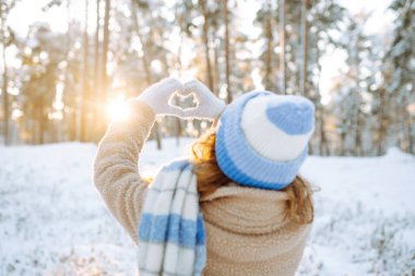Kar ormanının arka planında şapkalı ve eşarplı bir kadın ellerini kalp şeklinde tutuyor. Arkadan bak. Genç bir kadın güneşli bir kış gününün tadını çıkarıyor. Tatil konsepti, yaşam tarzı.