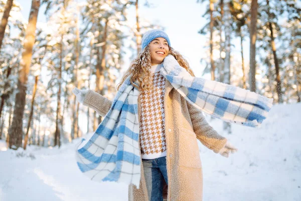 Playing with snow. Beautiful woman in a hat and scarf playing with snow, having fun in a snowy winter forest. Cheerful woman enjoying sunny frosty weather.