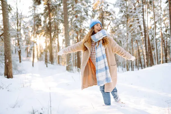 Playing with snow. Beautiful woman in a hat and scarf playing with snow, having fun in a snowy winter forest. Cheerful woman enjoying sunny frosty weather.